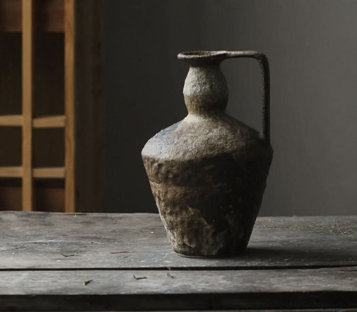 Textured, earth-toned wabi-sabi small ceramic vase with a handle, on a rustic wooden table.