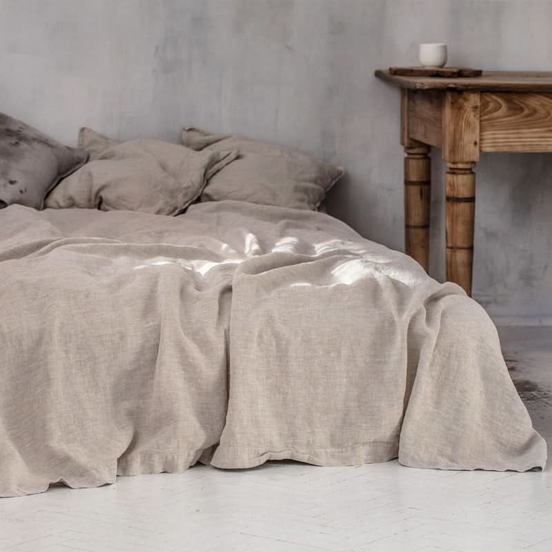 Natural linen bed sheets in beige, draped on a bed with linen pillows and a rustic wooden table.