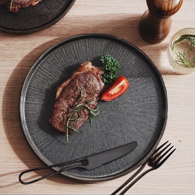Dark gray flat ceramic glazed plate with steak, tomato, herb, and black cutlery.