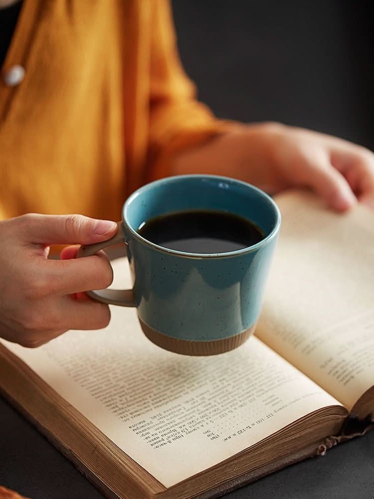 Glazed clay mug with a blue-green finish, held over an open book.