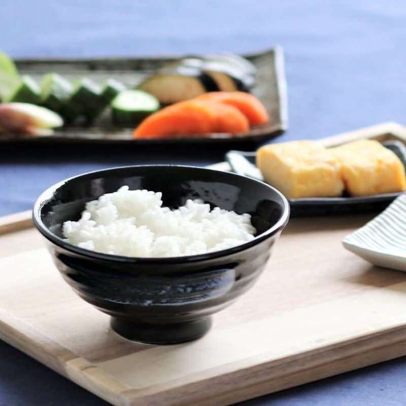 Black ceramic rice bowl with white rice, part of Japanese tableware set with tamagoyaki and vegetables.