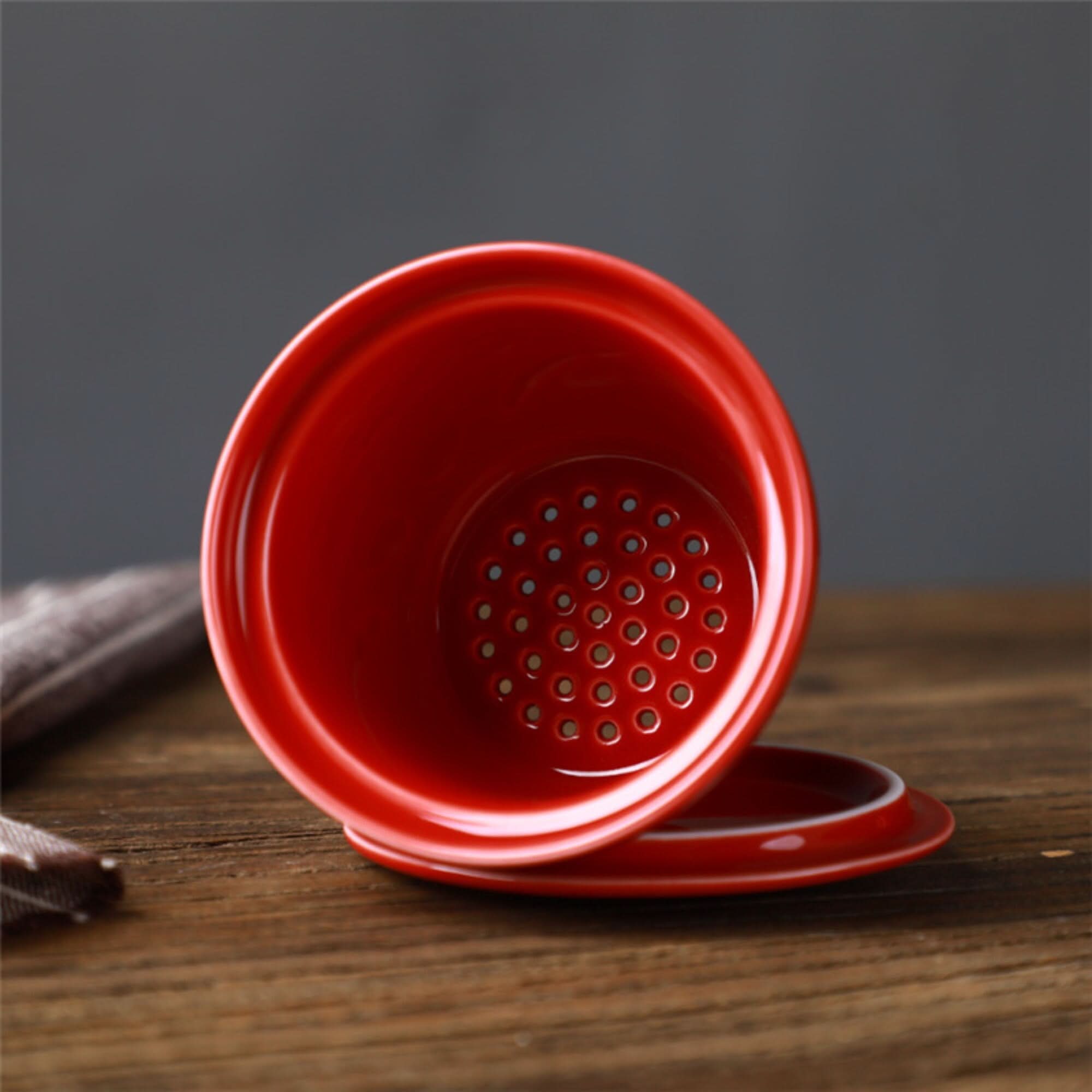 Red ceramic Japanese mug with lid and strainer on wooden table.