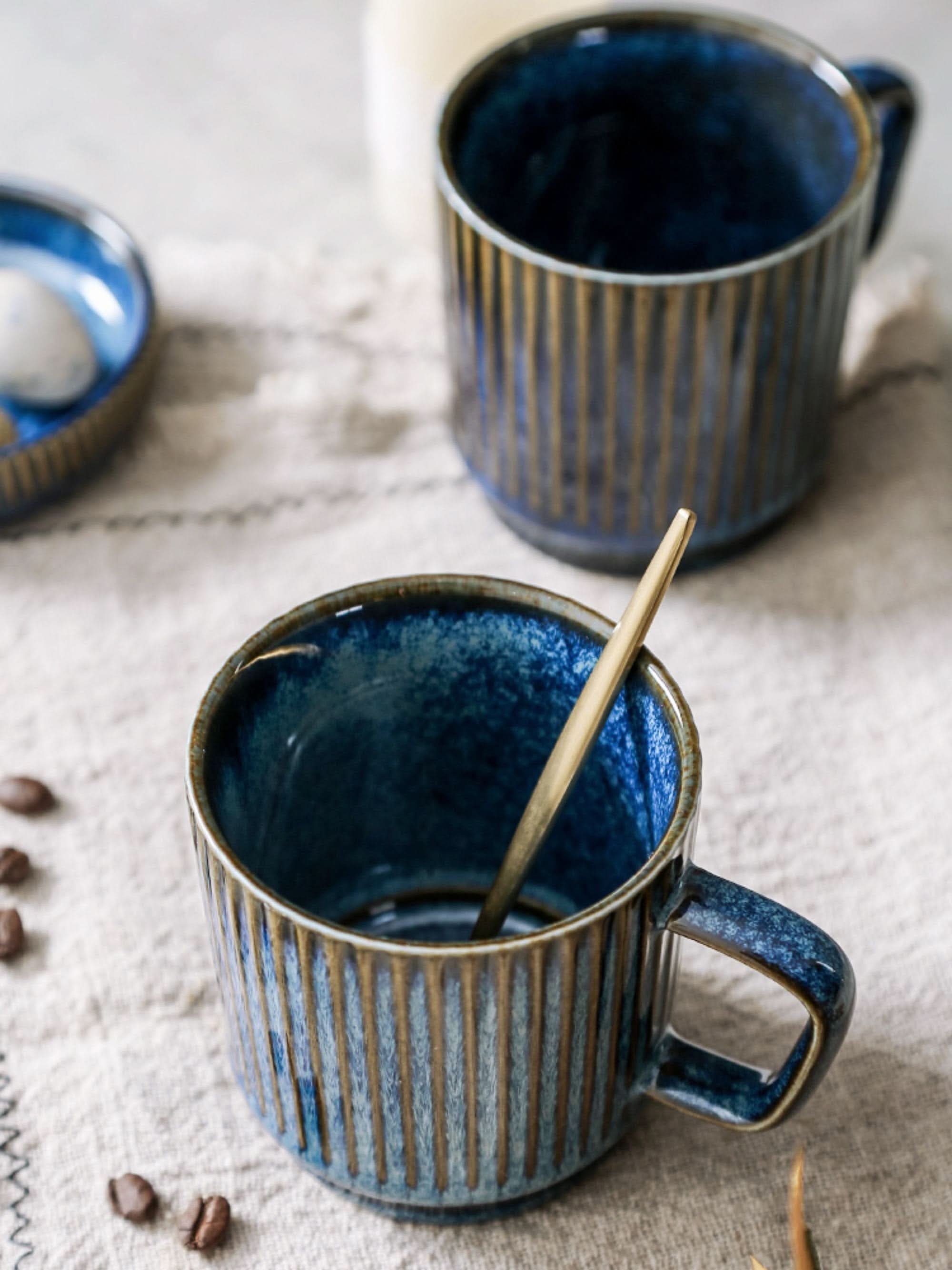 Blue glazed stoneware mug with vertical ribs, a gold spoon, and coffee beans on a textured cloth.