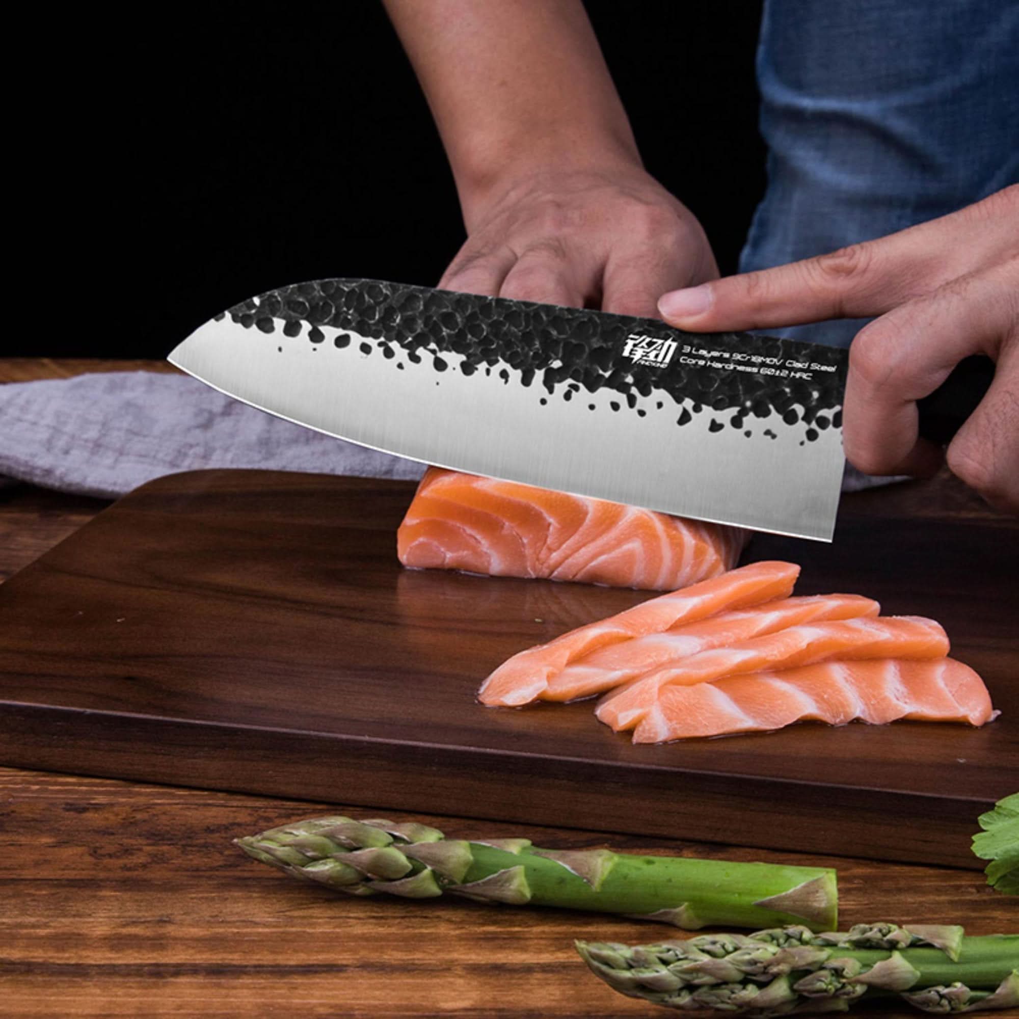 Black hammered Japanese Forged Knife slicing salmon on wood cutting board with asparagus.