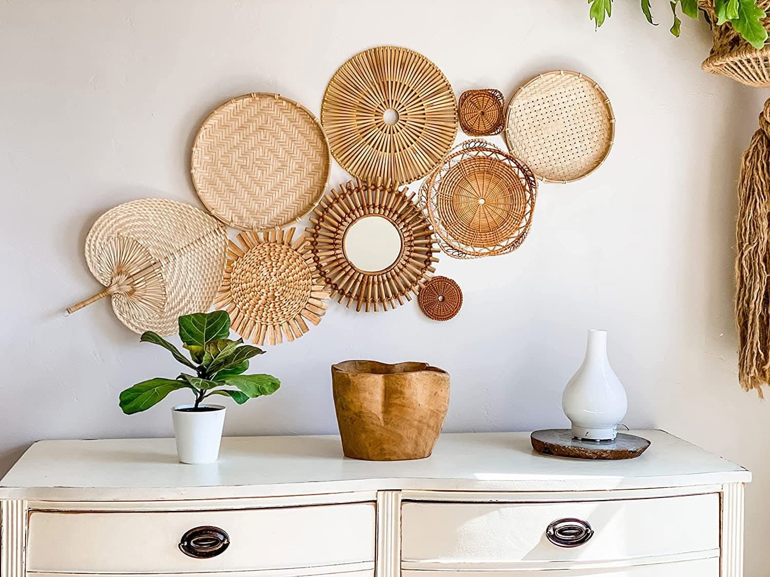 Handmade straw boho wall decoration set of woven natural rattan and bamboo discs, fans, and mirror on white wall.