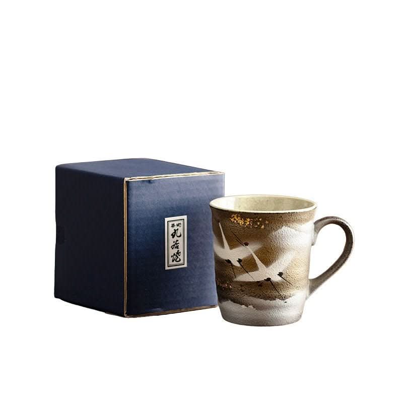 Kutani Burning Crane Mug Japanese-style, brown & white ceramic with gold accents, next to navy gift box.