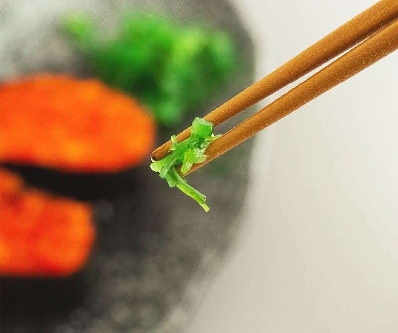 Wooden chopsticks holding green seaweed, with orange sushi in background. SUNLIFE tableware.