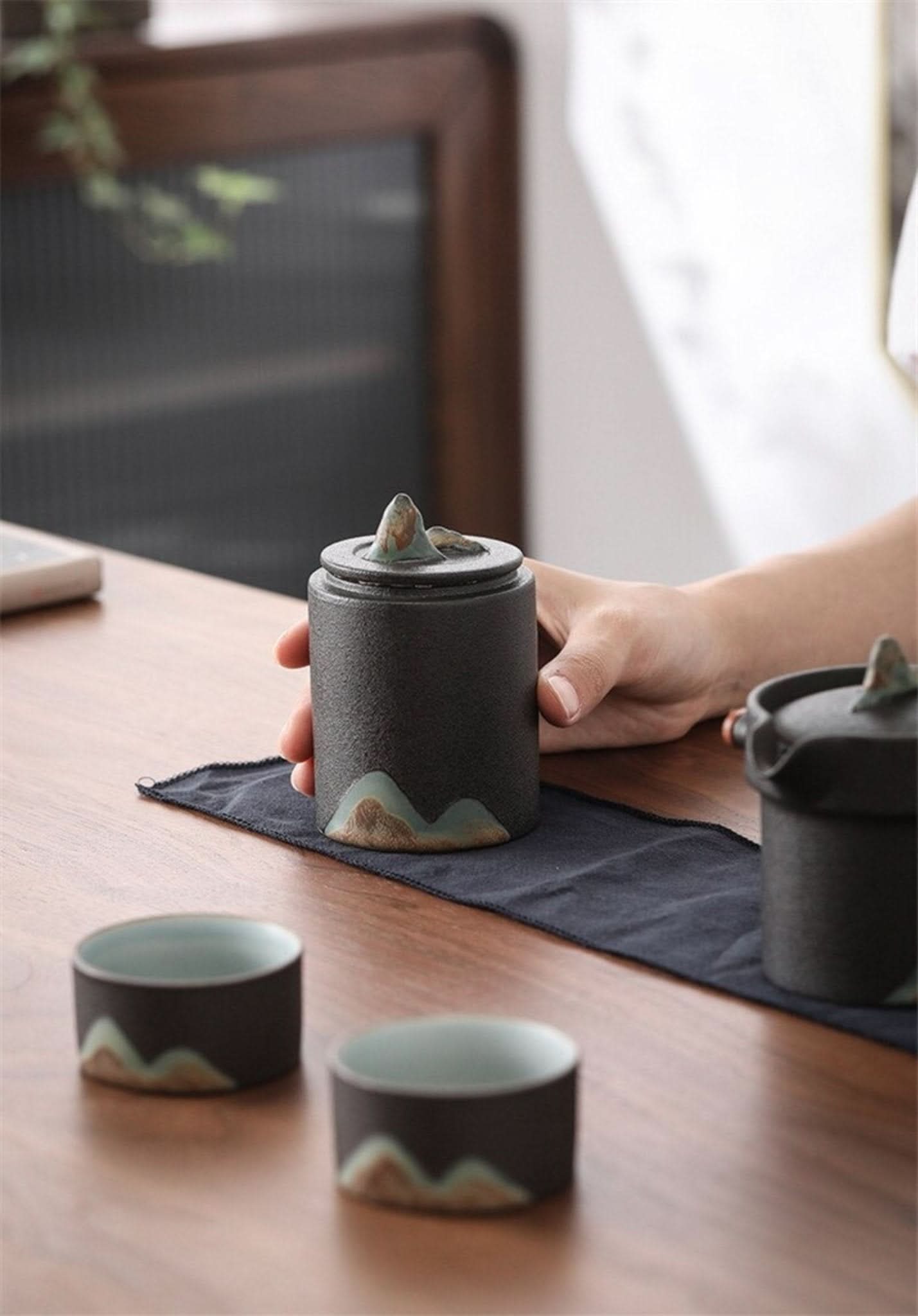 Black matte finish Ceramic Tea Canister with mountain motif, on wooden table with cups.