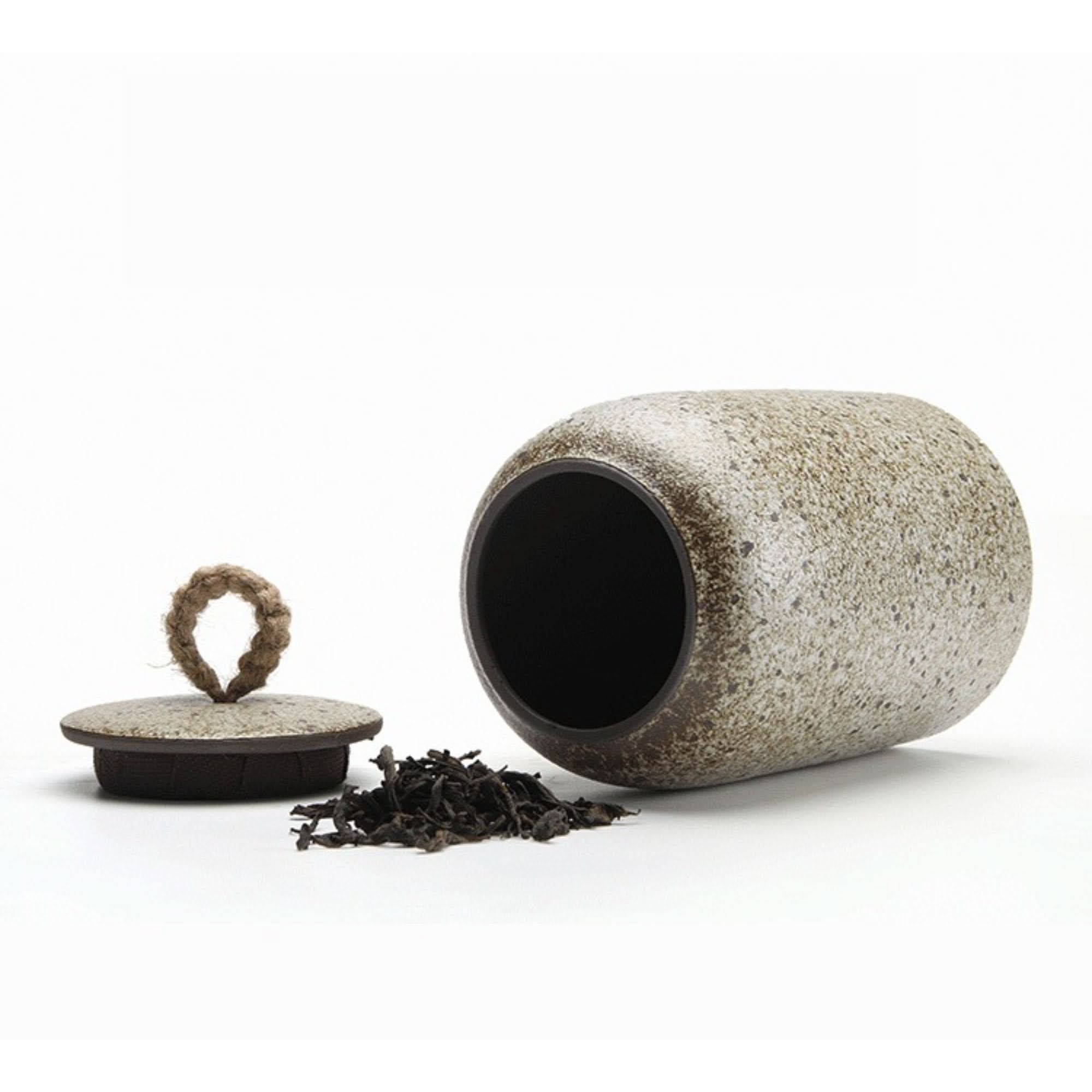 Speckled ceramic tea canister with a rope lid handle and loose tea leaves.