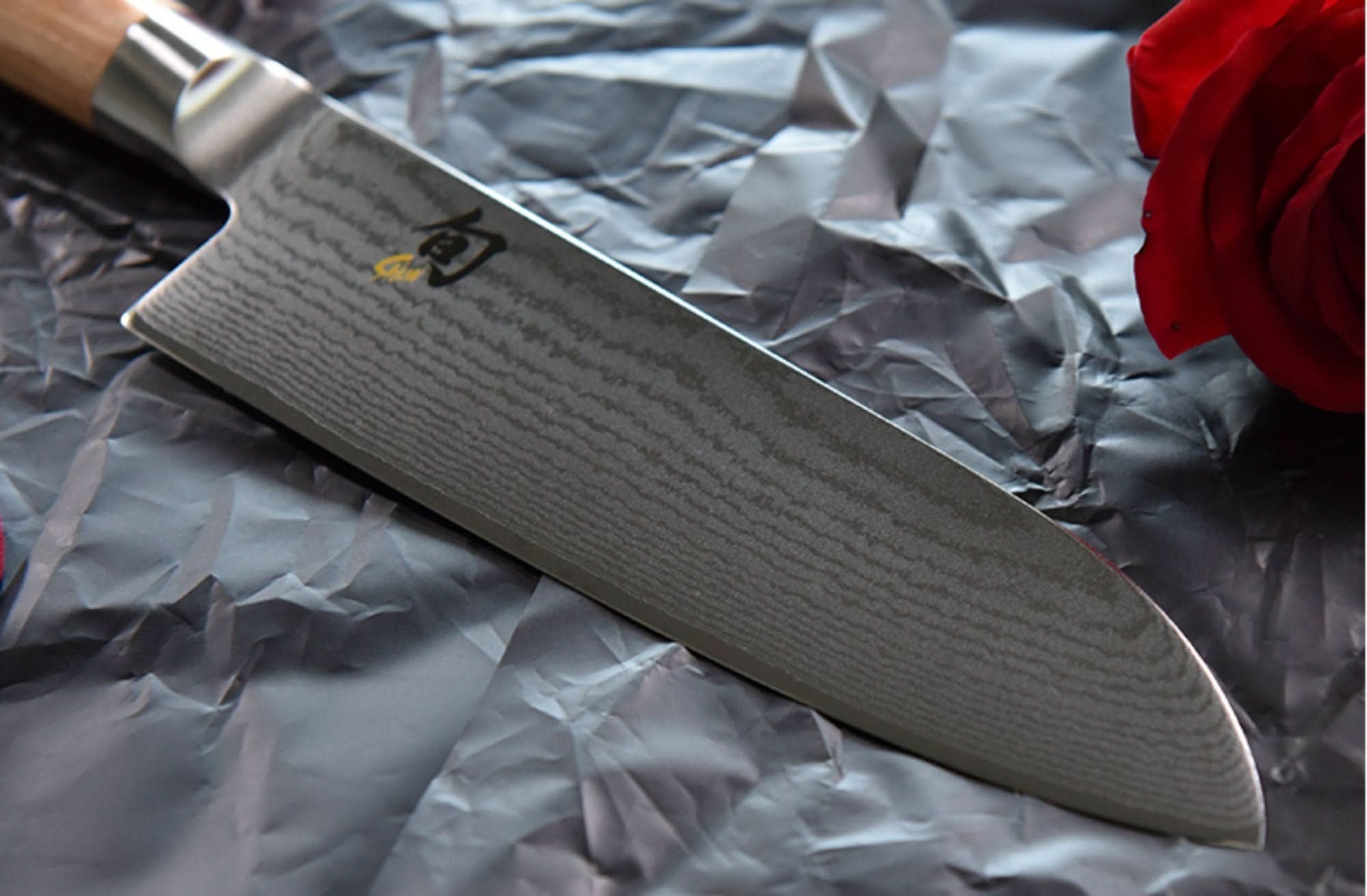 Japanese Santoku Damascus steel knife with wood handle on textured grey paper next to red rose.