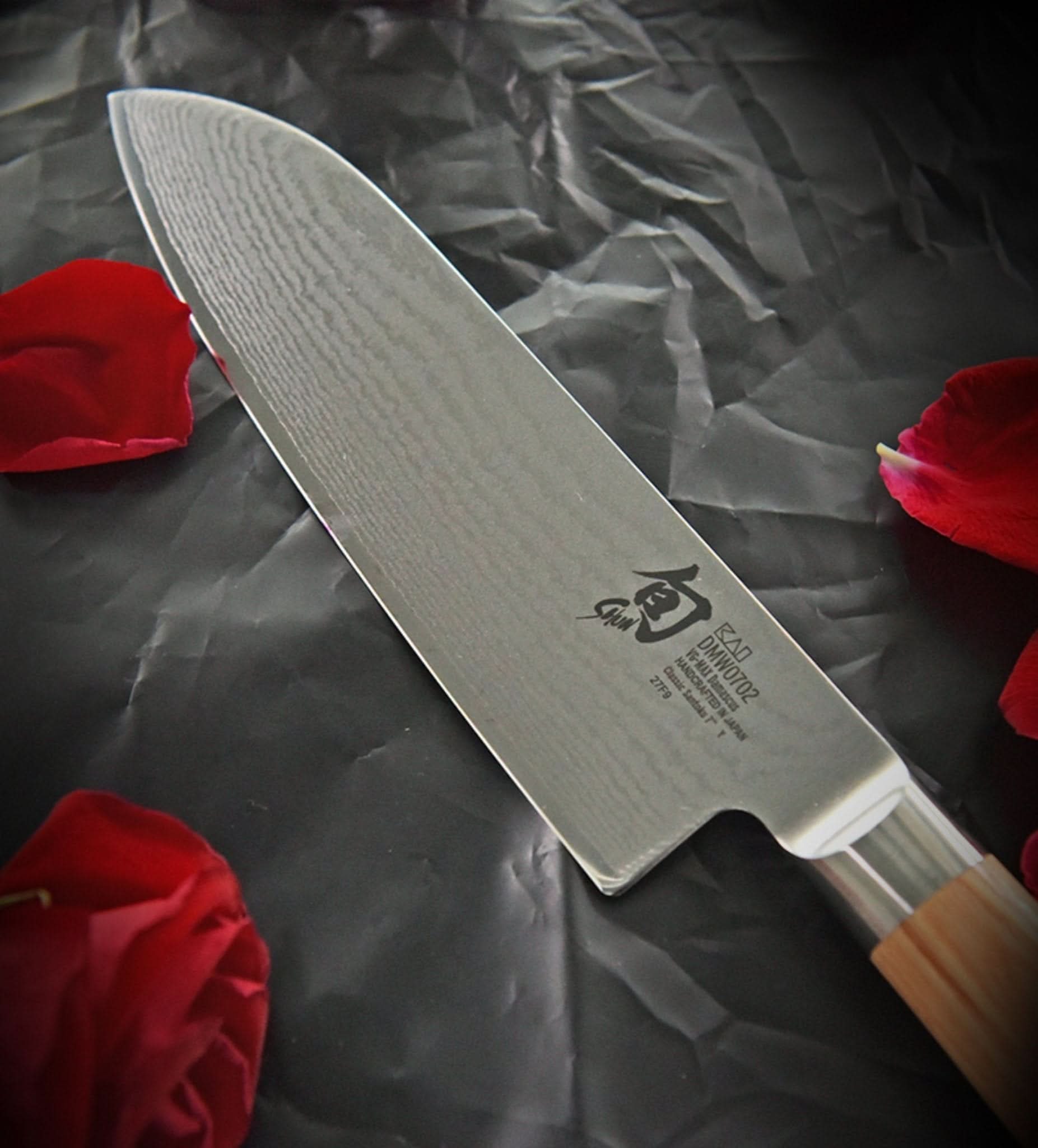 Damascus steel Santoku knife with wooden handle, set against dark fabric & red petals. Made in Japan.