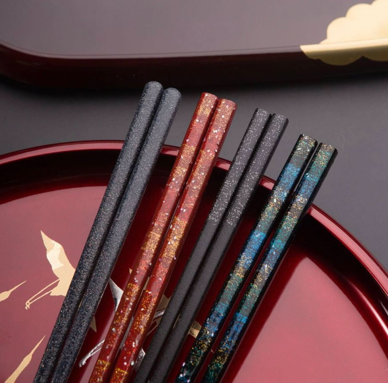 Japanese handmade wooden chopsticks in red, black, and blue with glittering accents on a dark red tray.