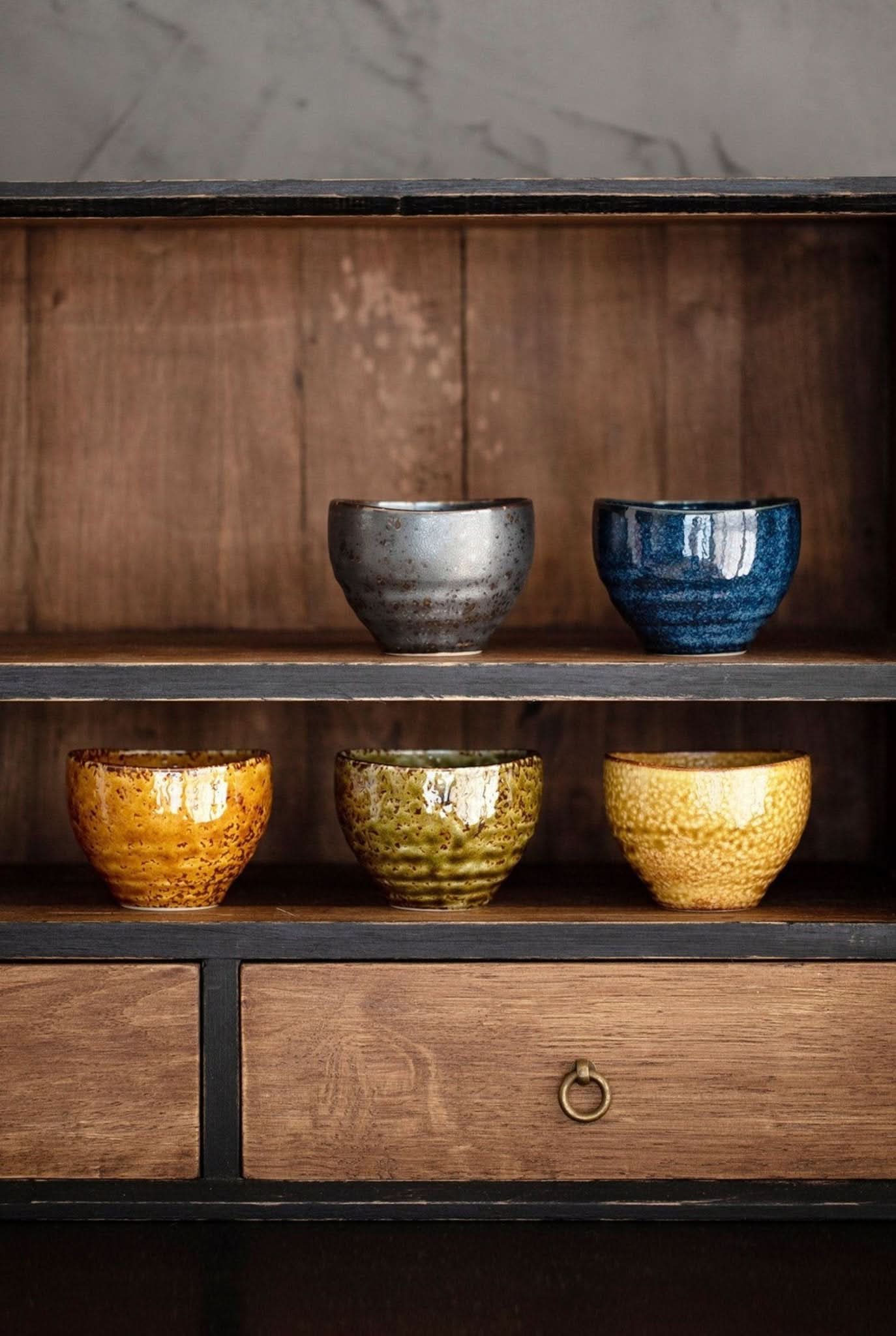 Japanese-Imported Glazed Tea Cup 5 Set: Five rustic ceramic tea cups in earthy tones, displayed on wooden shelves.
