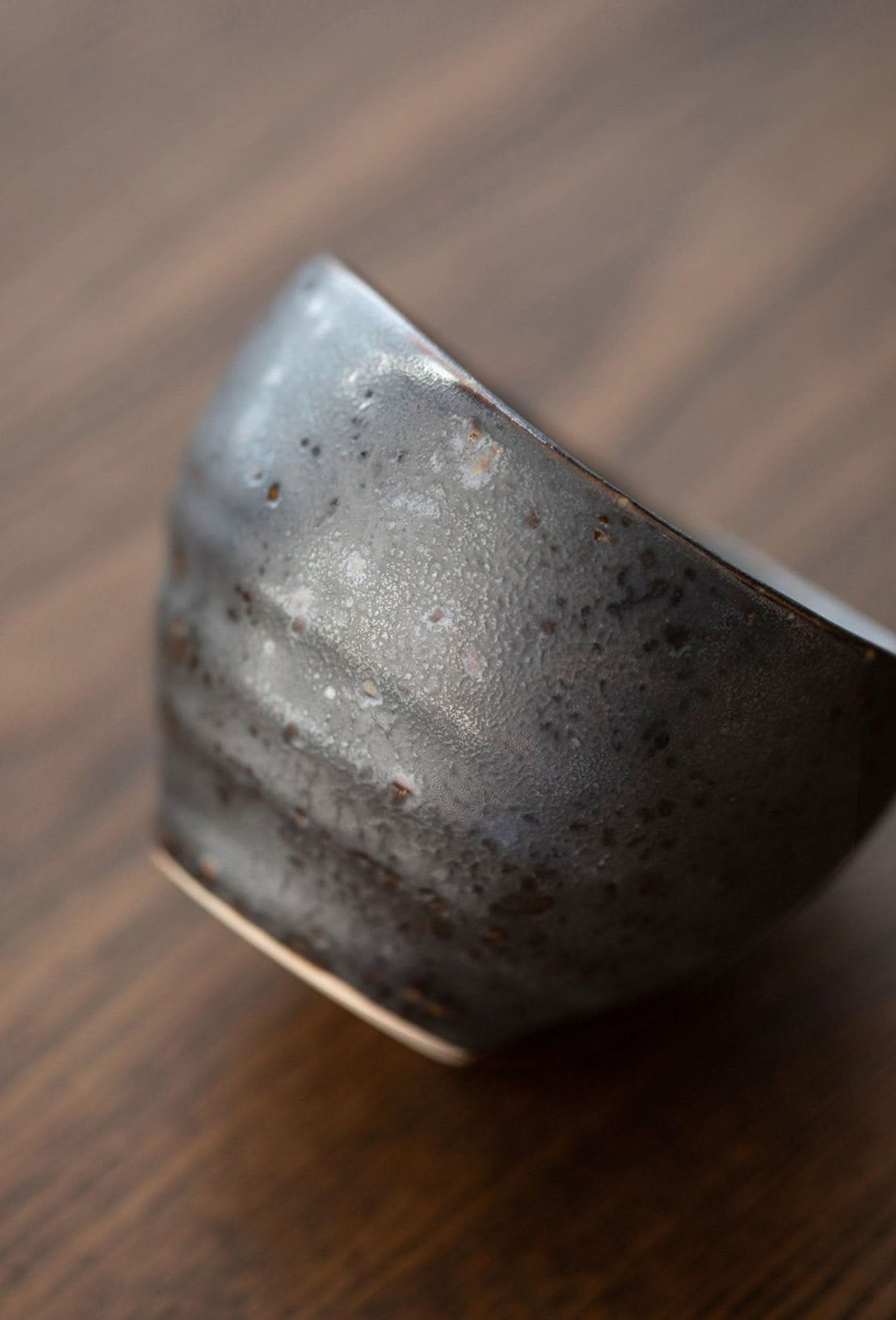 Japanese-imported glazed tea cup, dark ceramic with speckled texture on wooden surface.