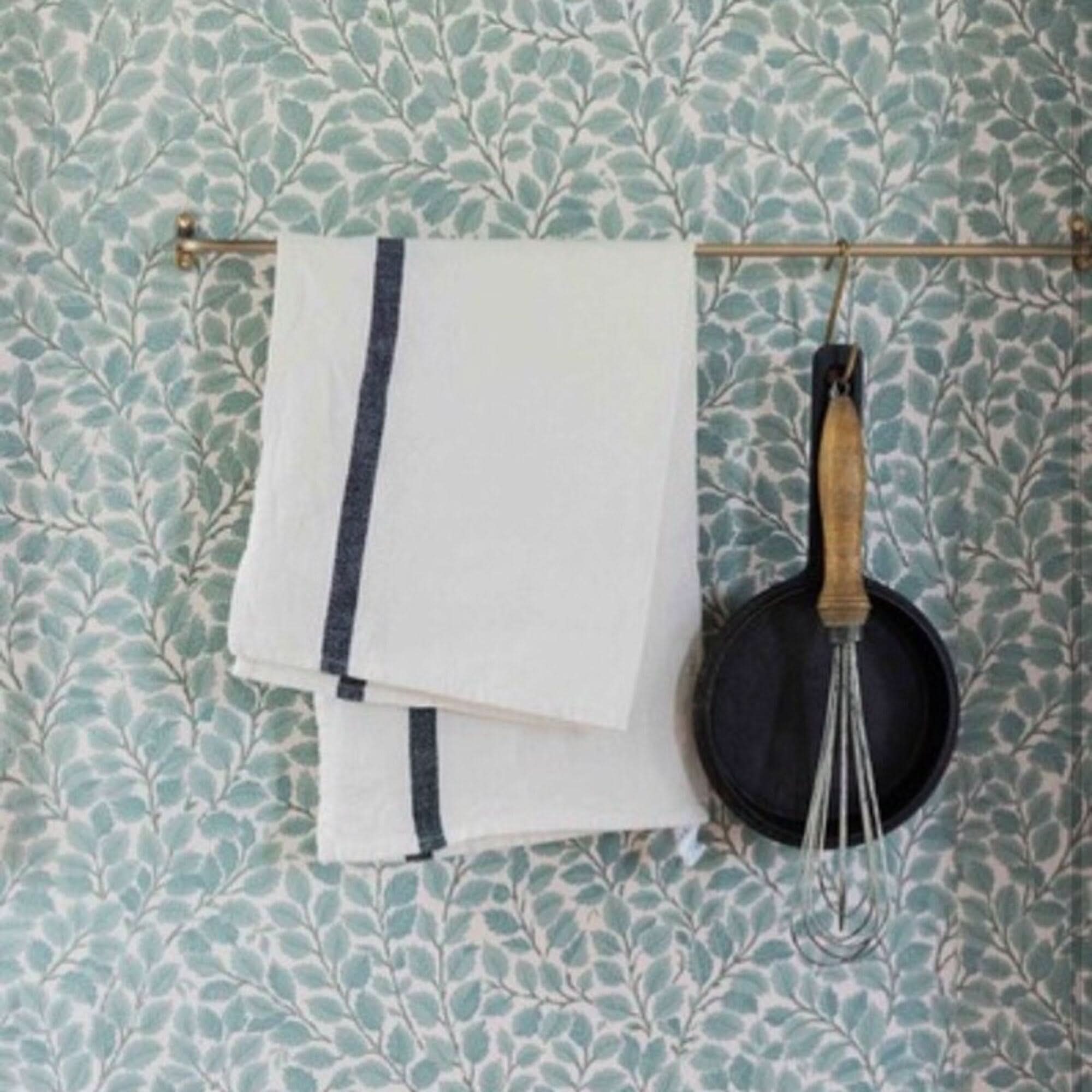 White Japanese linen tea towels with navy stripes, hanging on a brass rod against patterned wallpaper.