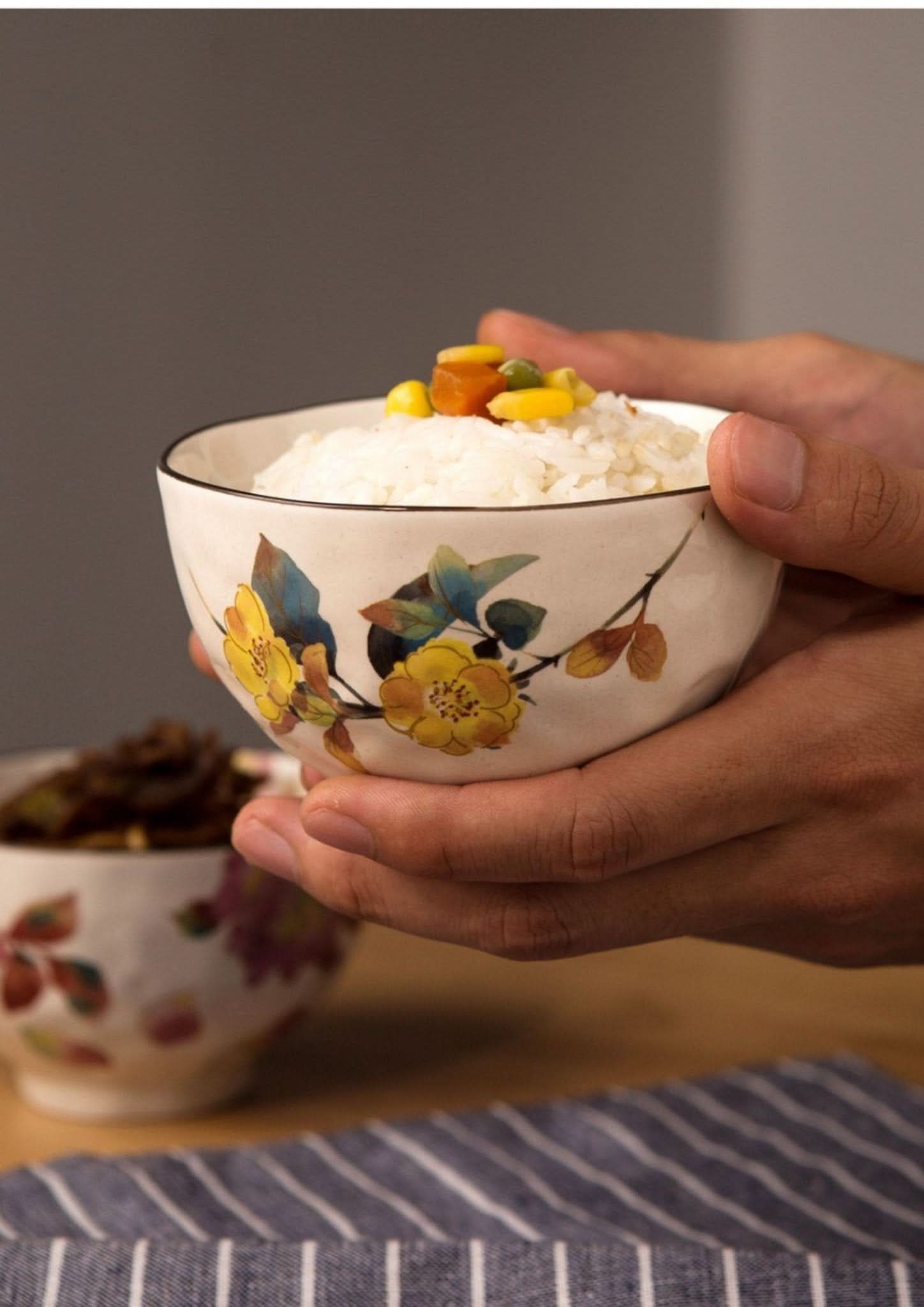 Hands hold a floral ceramic rice bowl set made in Japan, filled with rice and vegetables.