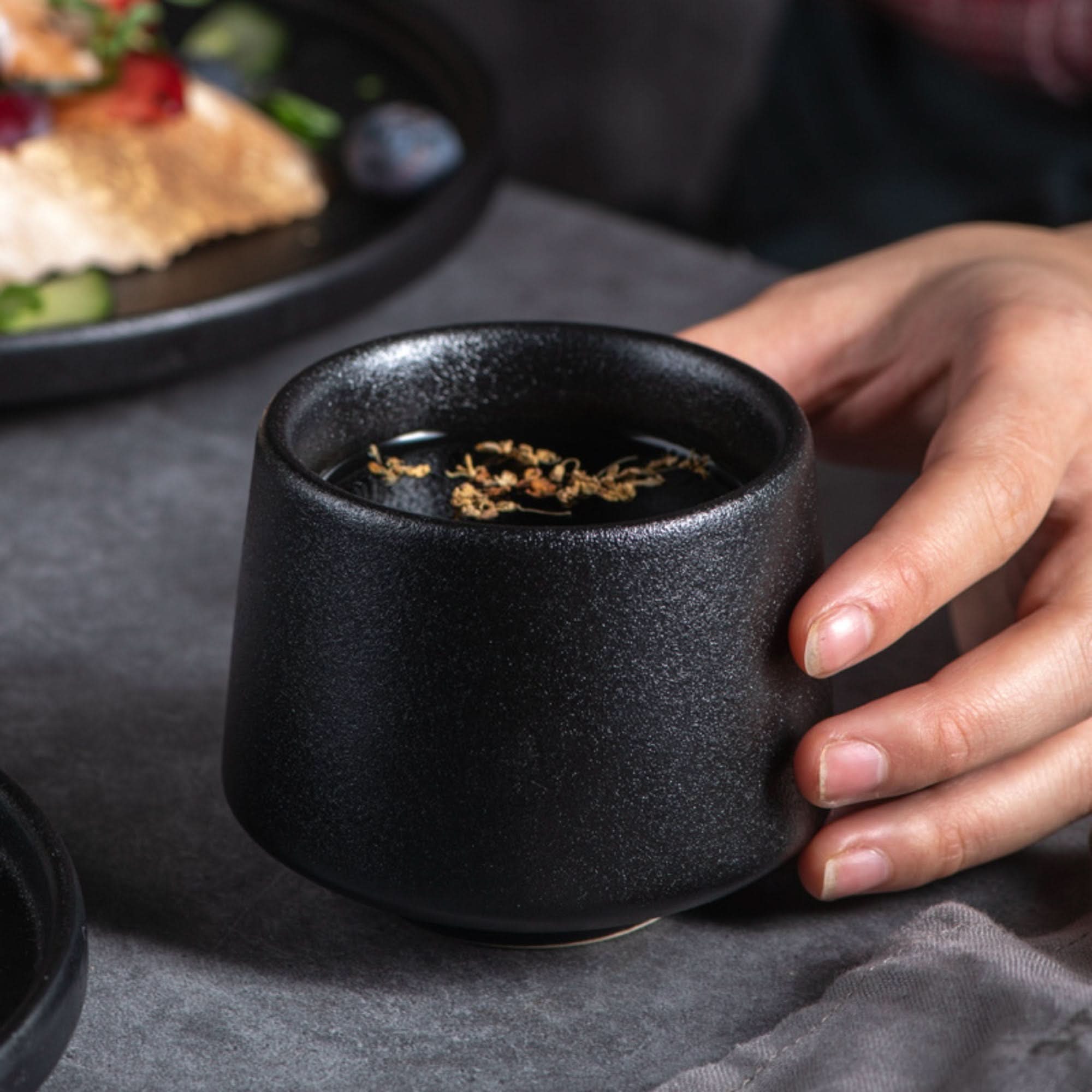 A hand holds a textured, gray-toned Japanese tea cup filled with dark tea and herbs.