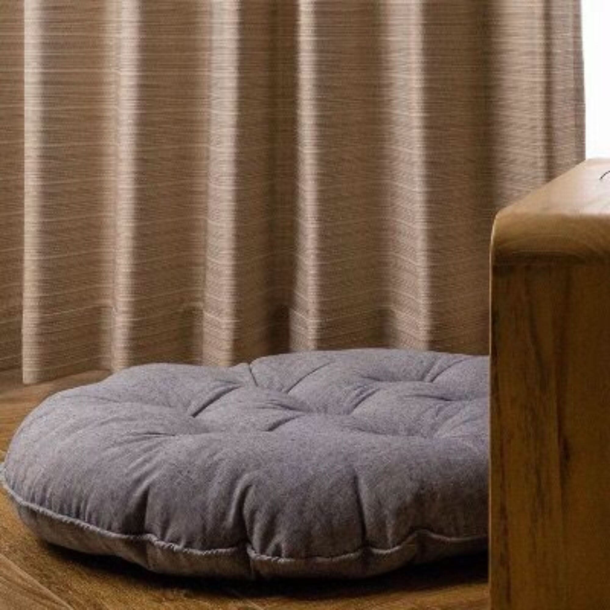 Gray linen meditation cushion, tufted design, sits on wooden floor near beige curtains.