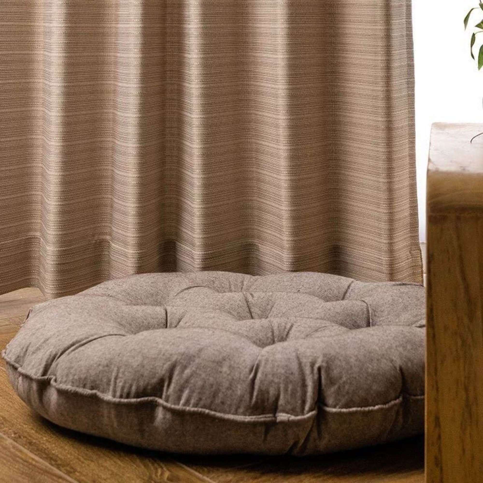 Taupe linen meditation cushion on wooden floor, next to beige textured curtains.