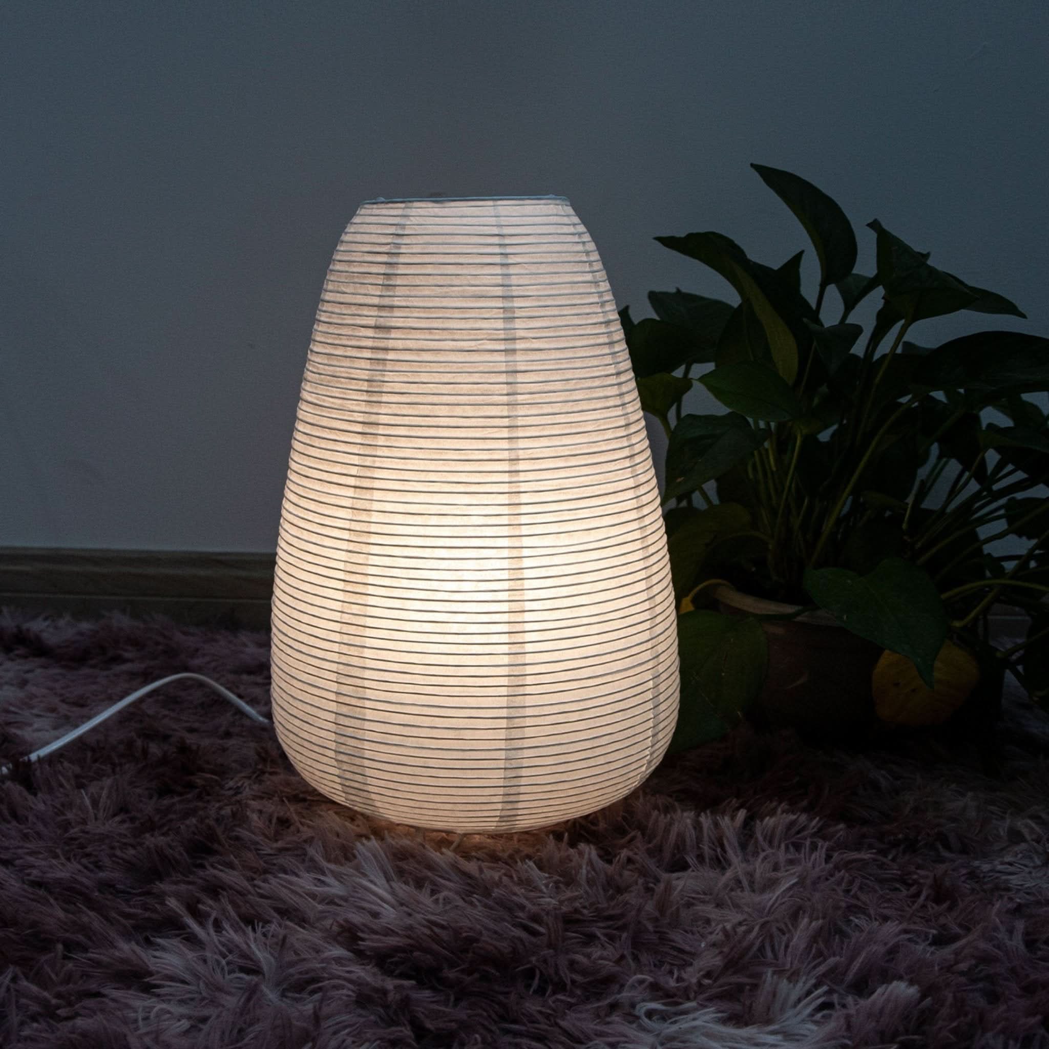 Paper Table Lamp Elongated emits soft light on a textured rug, with a plant in the background.