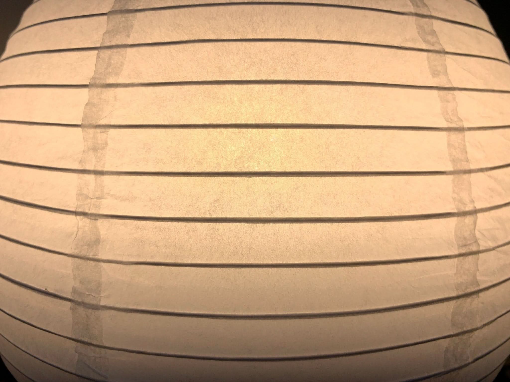 Close-up of a glowing white paper table lamp with horizontal lines and a vertical seam detail.