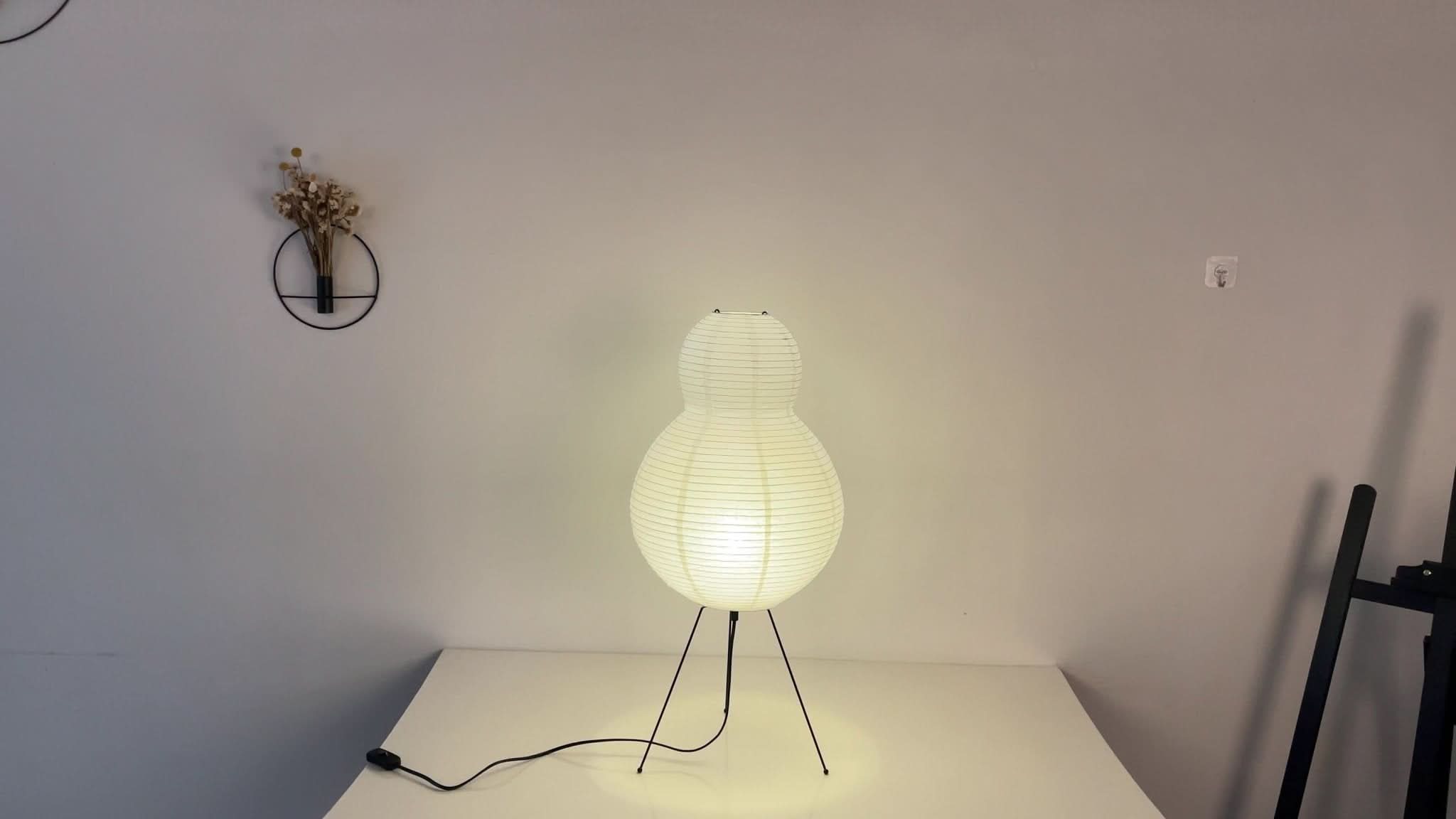 White paper floor lamp with pear shape, on black tripod legs, on white surface.