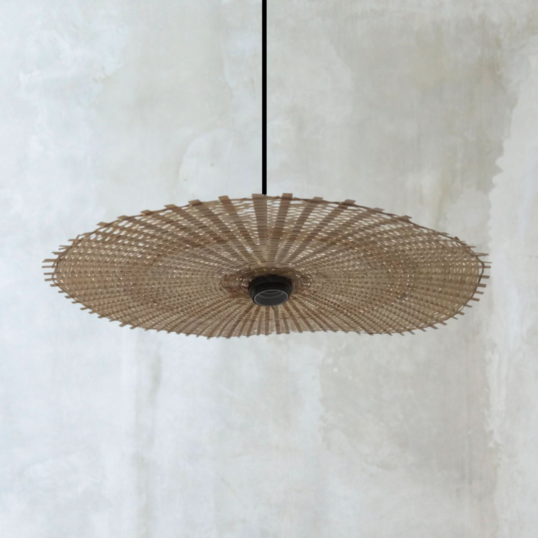 Natural rattan lamp shade with hat shape, hanging from a black cord against a textured white wall.