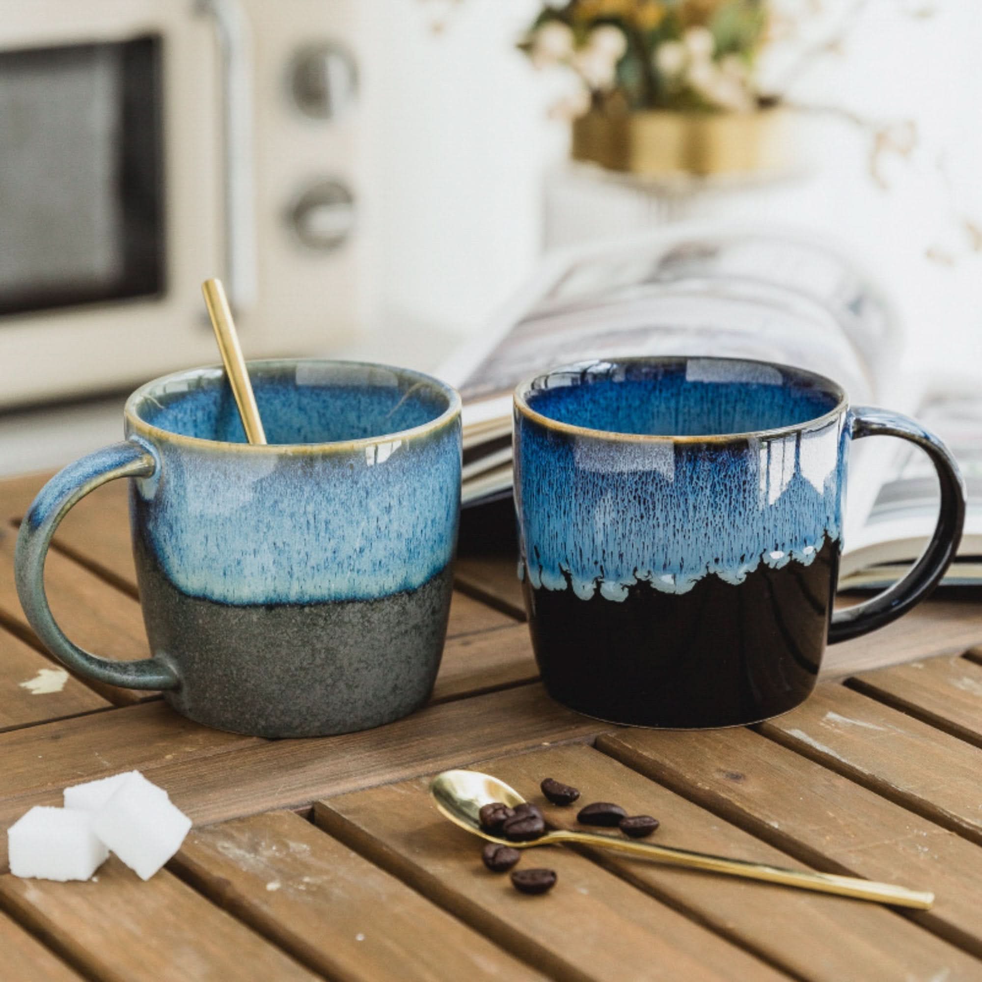 Two reactive glazed mugs, blue and gray ceramic, with coffee beans and sugar cubes on a wooden table.