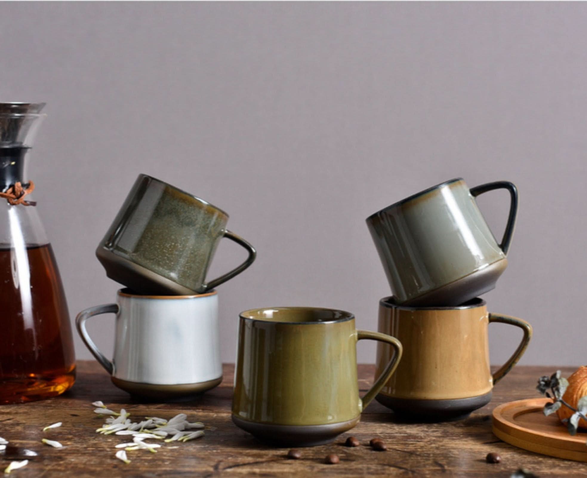 Earth-toned glazed ceramic mugs stacked on a wooden table, with a coffee carafe.