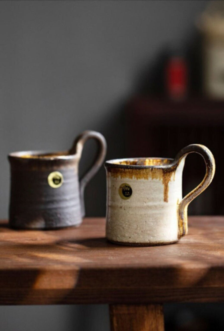 Two Shiraku-yaki stoneware mugs, one dark brown, one cream, on a wooden table.