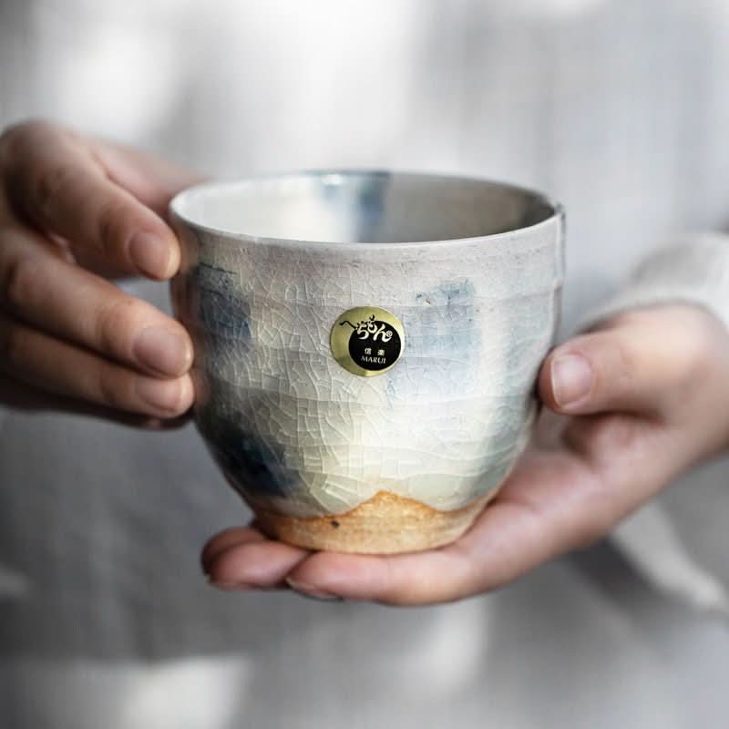 Hands hold Shiraku-Yaki tea cup, white ceramic with blue glaze & crackled finish, Japanese handmade.