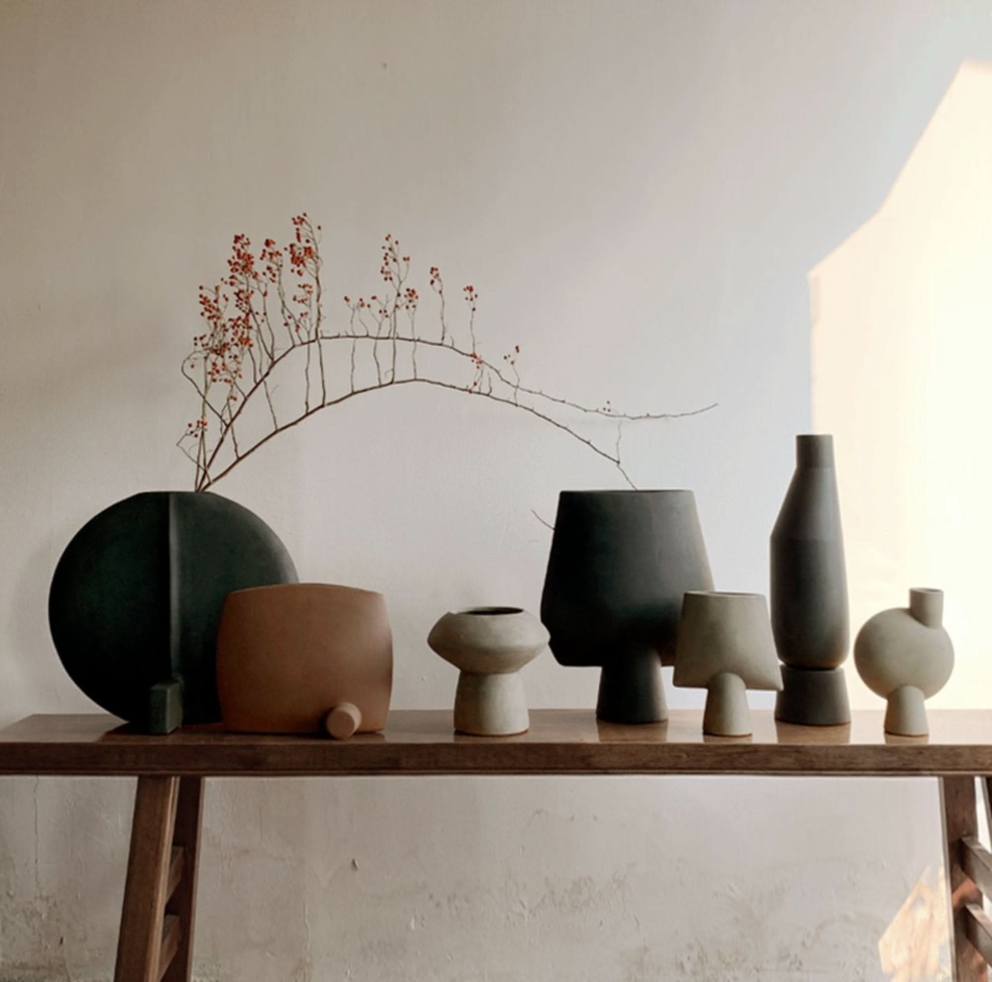 Small ceramic vases in black, gray, and brown, arranged on a wooden shelf with dried branches.