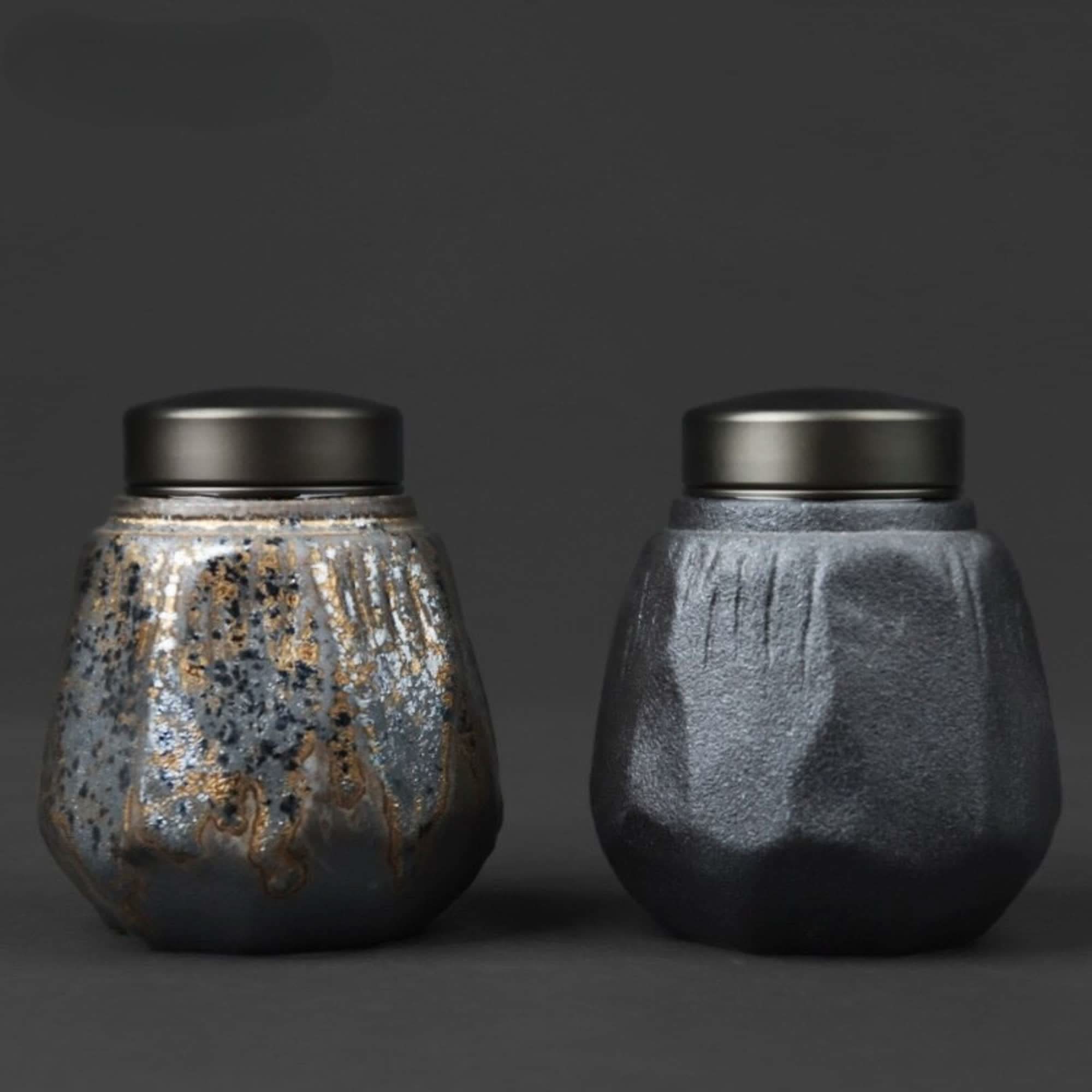 Two small stoneware sealed tea cans, one with a textured metallic glaze and gold flecks, on a dark background.
