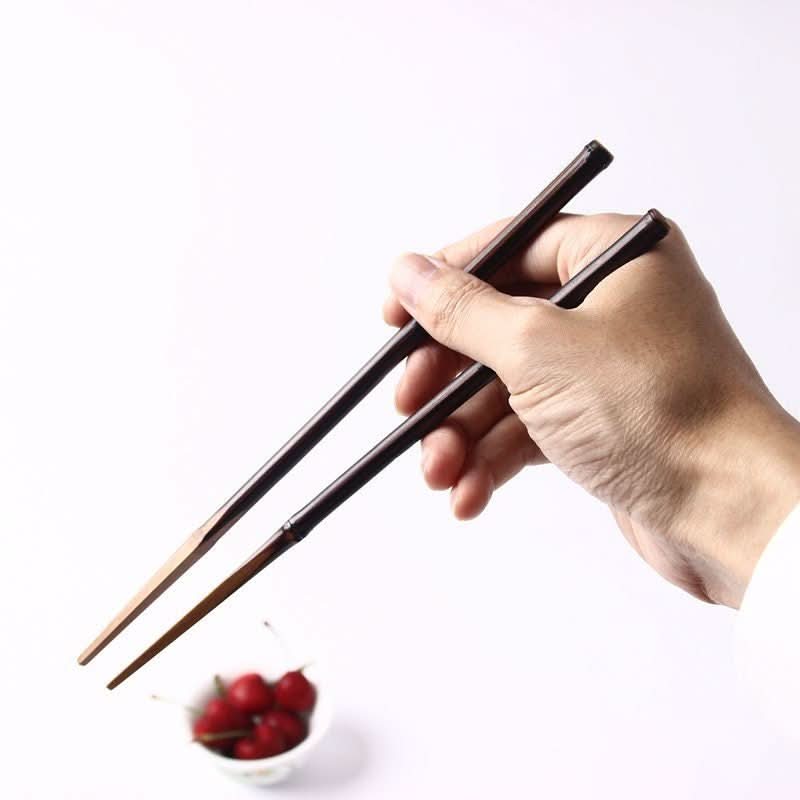 Solid bamboo chopsticks with glossy finish, held by a hand over a bowl of cherries.