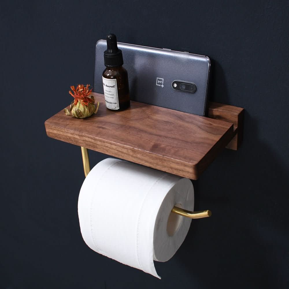 Solid wood and gold toilet paper holder with shelf, featuring a phone, bottle, and decorative item on the shelf.