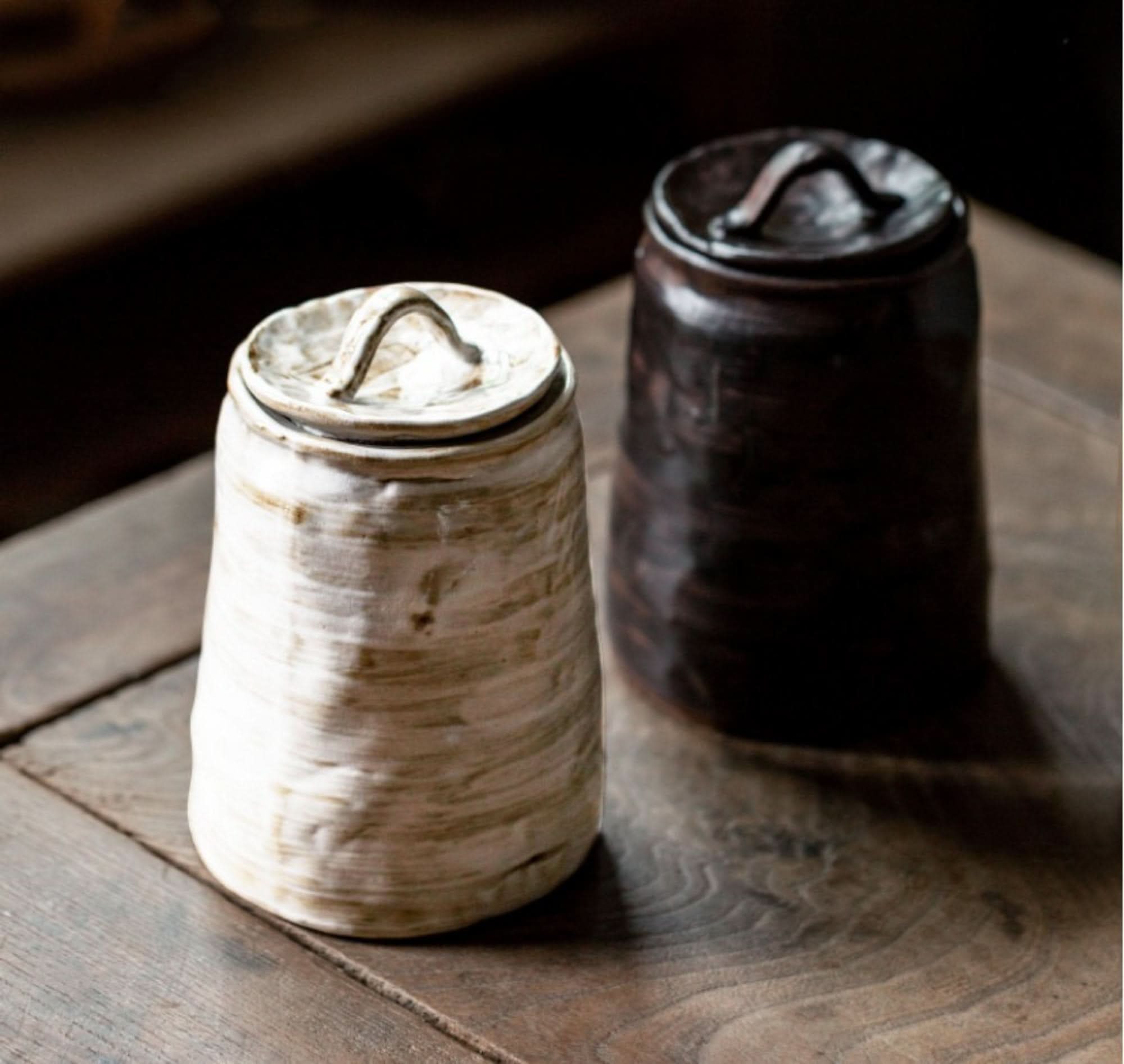 Two stoneware tea canisters, one white, one dark, with handles on wooden surface.