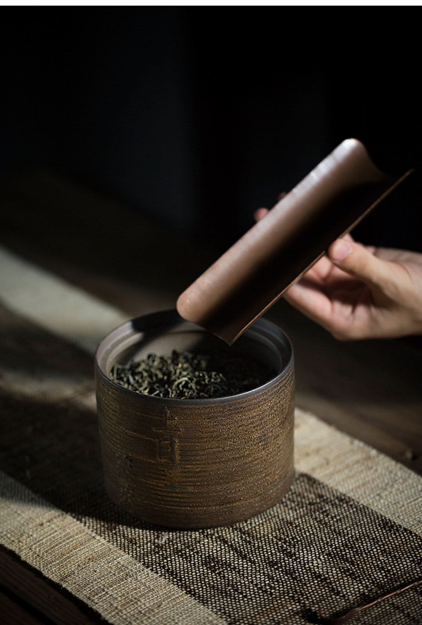 Japanese style ceramic jar, textured brown, filled with tea leaves, lid being placed by hand.