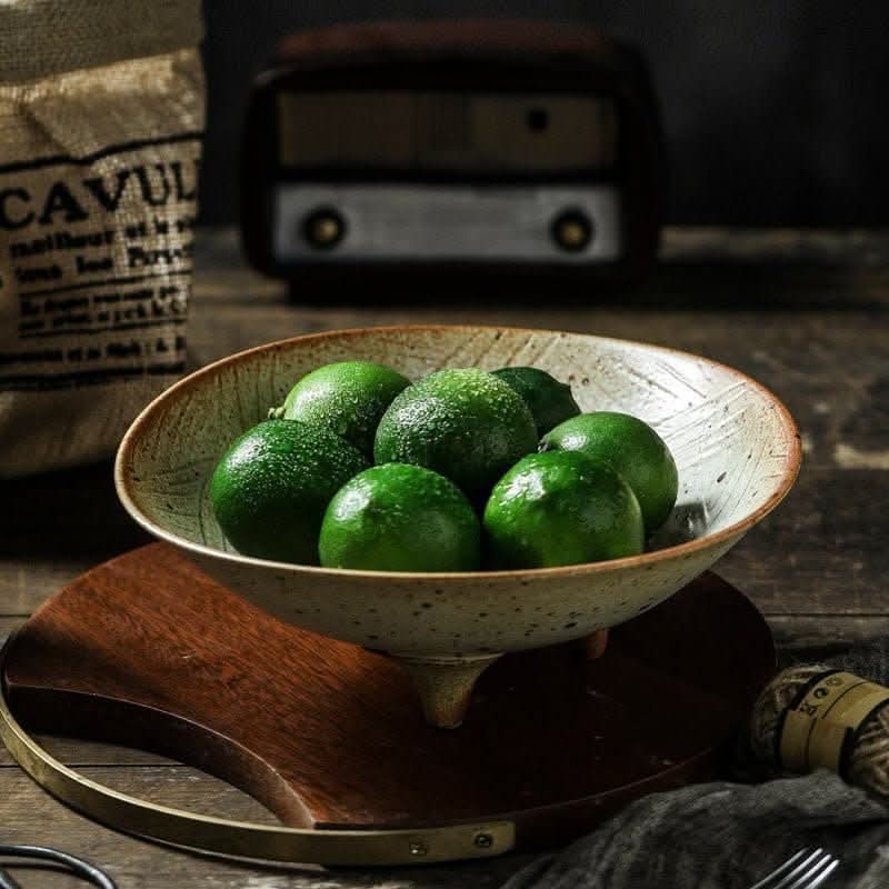 Tripod stoneware fruit plate holding fresh green limes on a wooden stand.