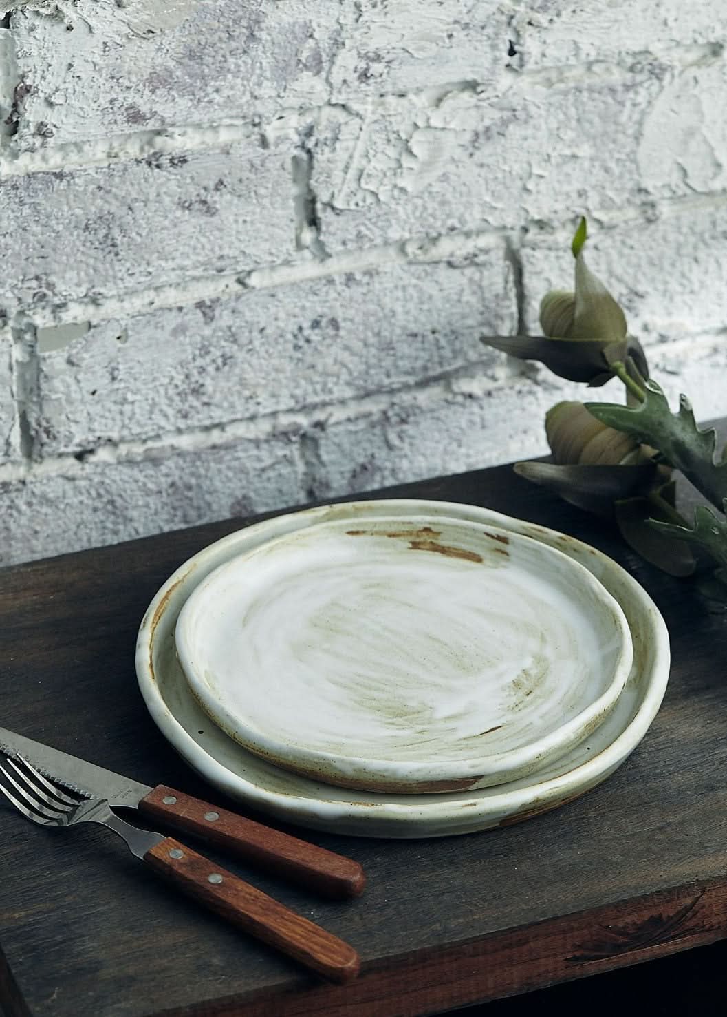 Stoneware plates with white glaze and earthy brown accents on a wooden table, with brick background.