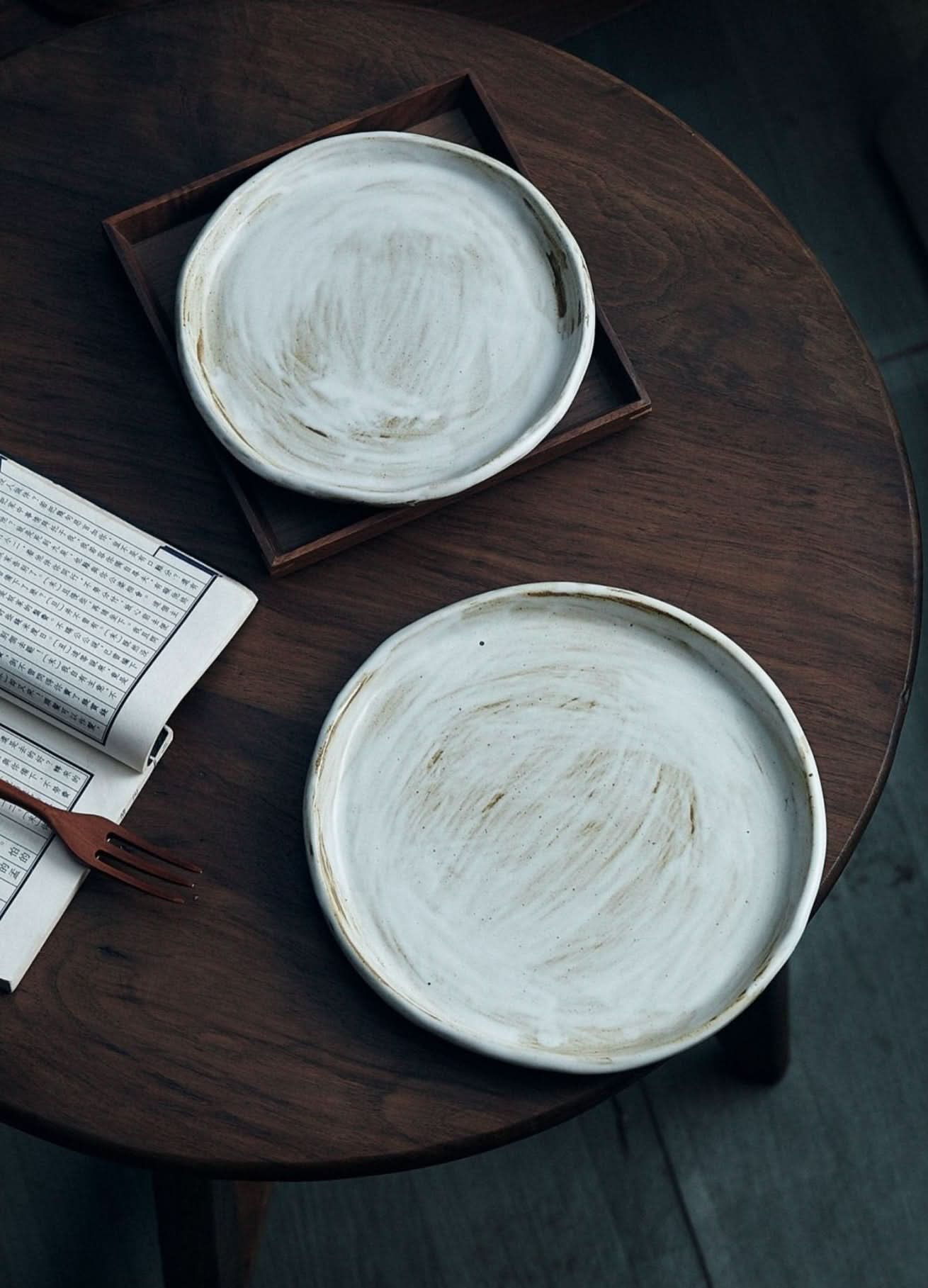 Two stoneware plates with white glaze, one on a wood tray, on a dark wood table.
