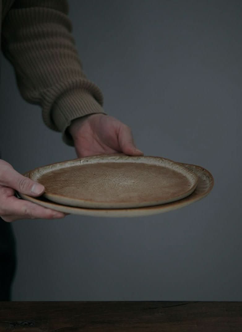 Unglazed large irregular coarse pottery flat plate held by hands against a neutral background.