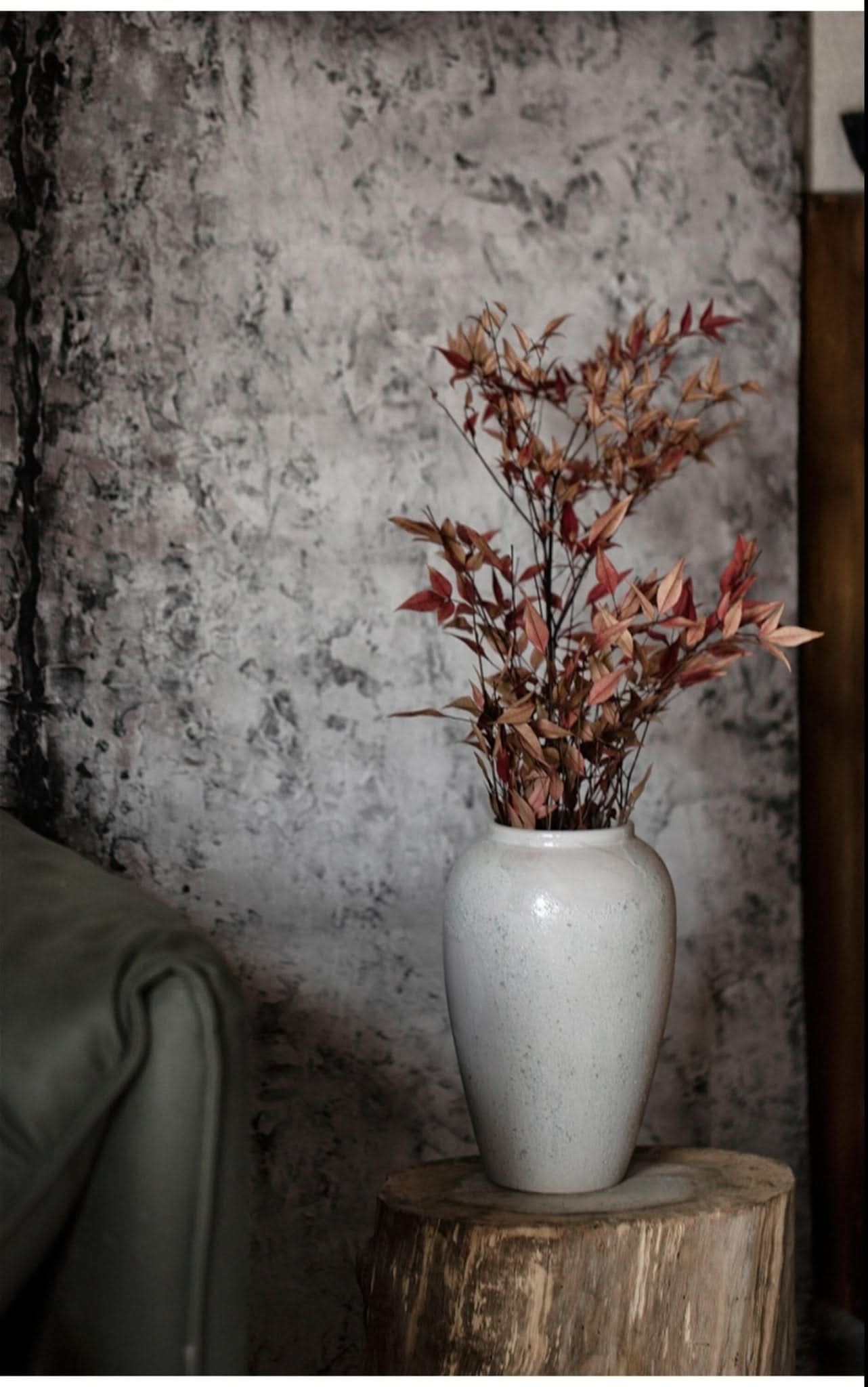 White ceramic vase with mixed texture holding autumn branches on rustic wood, grey textured background.