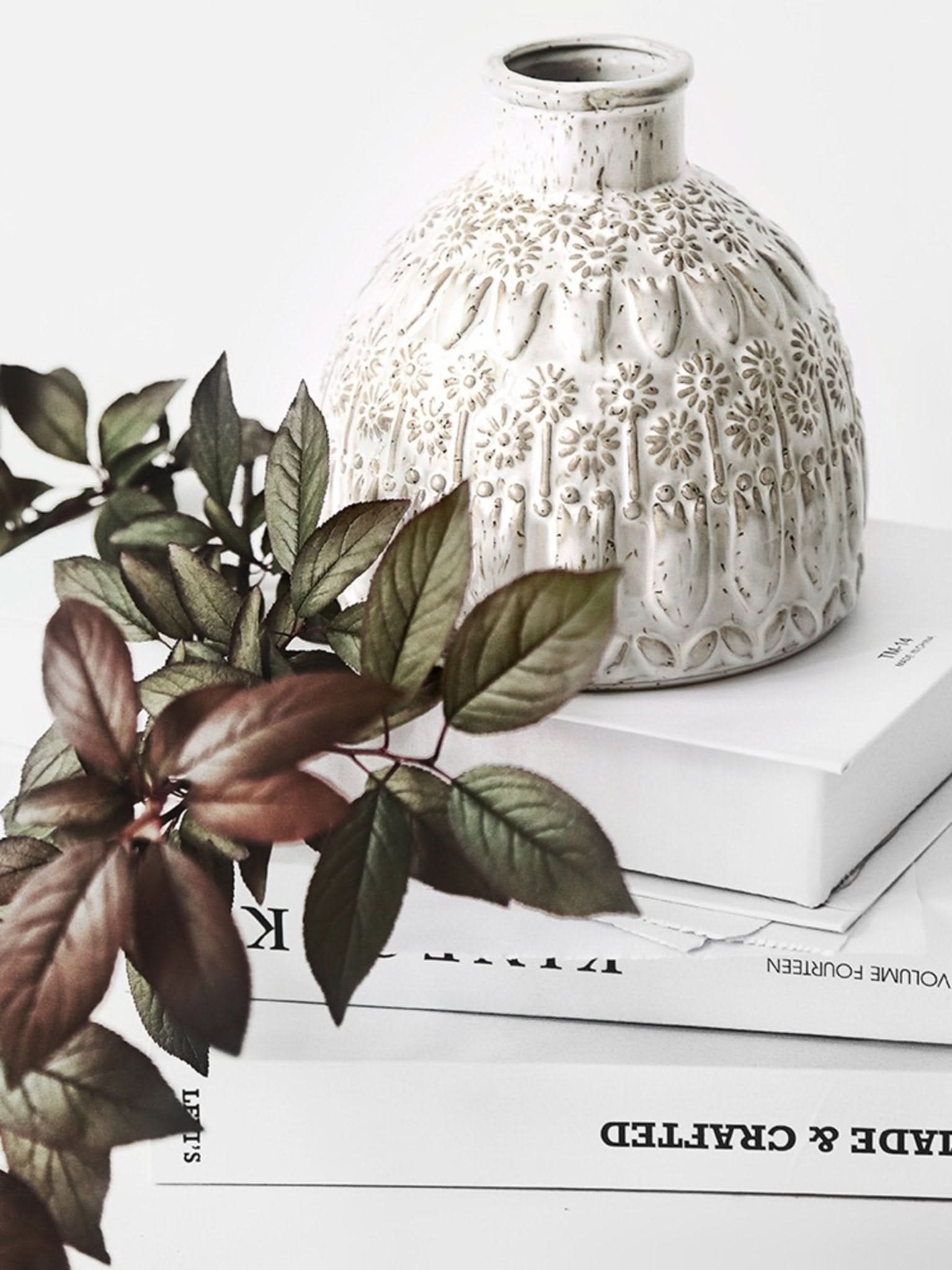 White ceramic small vase with textured floral design, next to green leaves on books.