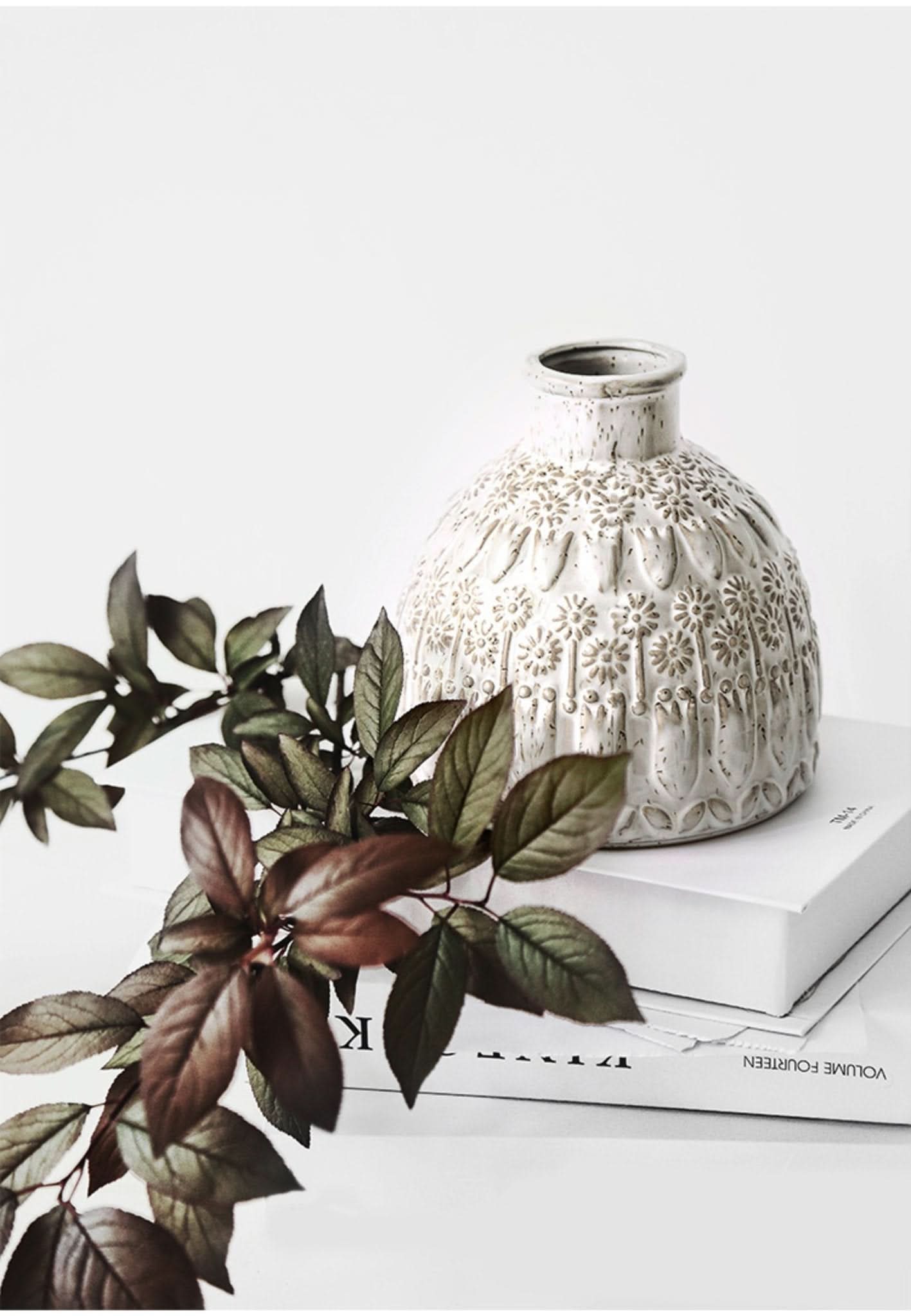 White ceramic small vase with embossed floral detail, styled with dark foliage on white books.